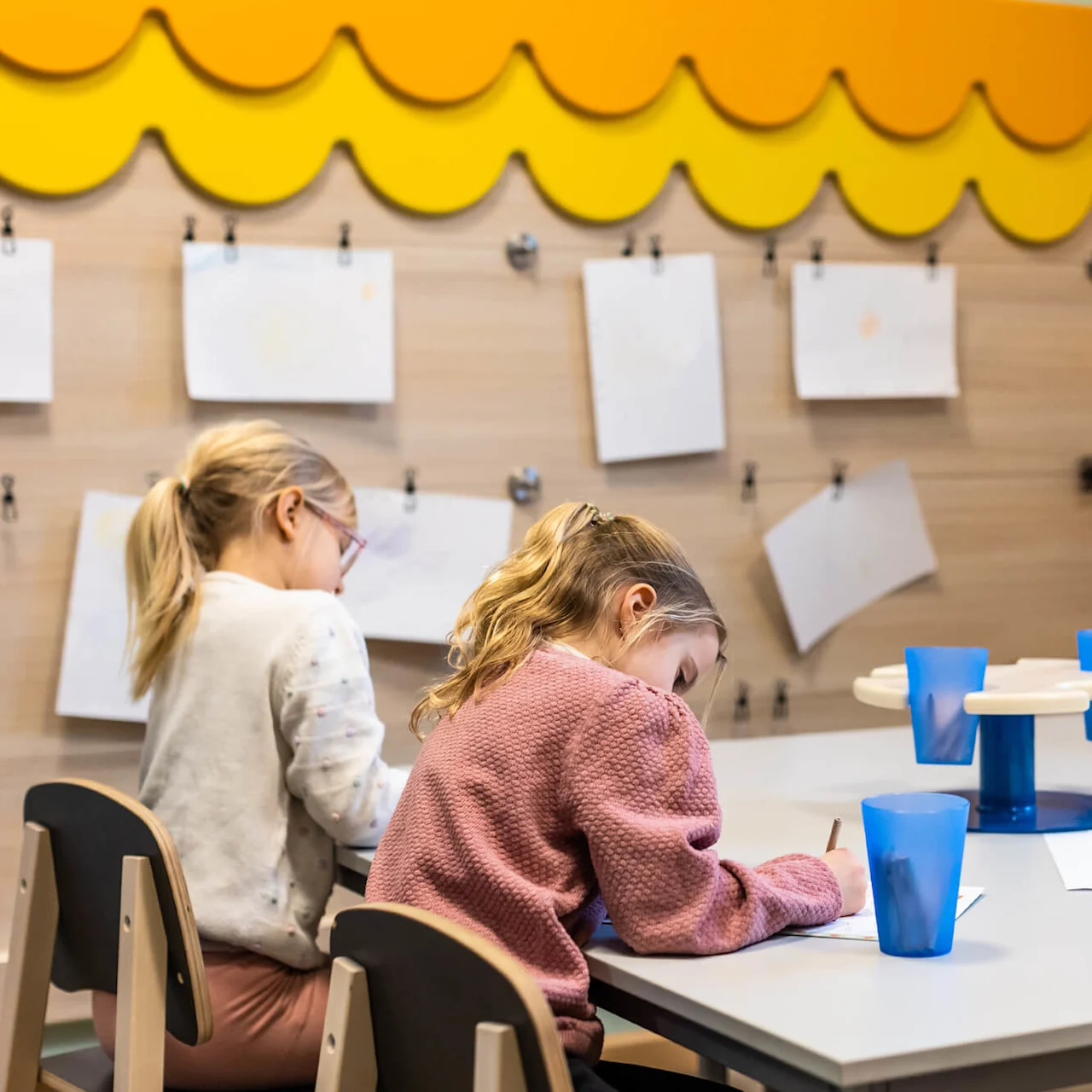 Børn tegner med farveblyanter ved et lille bord i et klasseværelse. Papirer hænger på væggen bag dem, og blå kopper er på bordet.