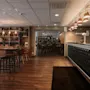 A cozy restaurant and bar area with warm lighting, wooden floors, a long bar, and bookshelves filled with books and plants.