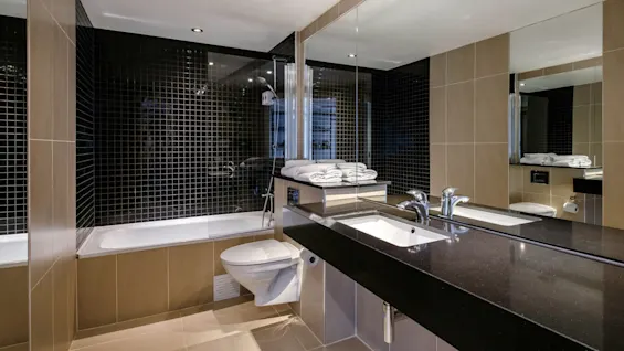 Et moderne bad har en vegg med svarte fliser, et badekar med sammenbrettede hvite håndklær, en vegghengt toalett og en elegant svart benkeplate med stort speil.