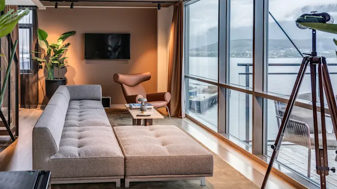 A modern living room features a gray sectional sofa beside a large window overlooking water. A cozy armchair, flat-screen TV, and telescope add comfort and functionality.