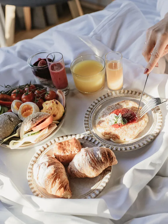 En person i morgonrock njuter av en härlig frukost i sängen med olika rätter och drycker, badad i naturligt ljus.
