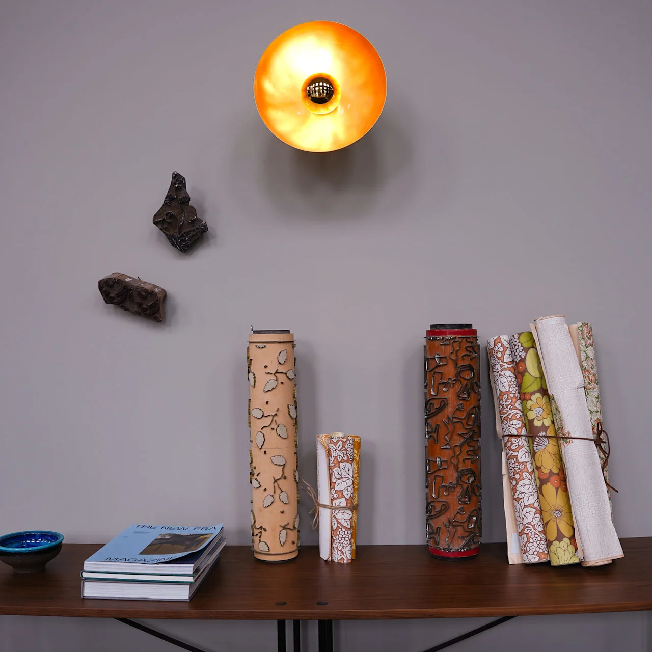 A wooden table with books, decorative scrolls, and a bowl, under a lit wall lamp; artworks on the wall.