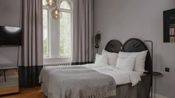 En omsorgsfullt bäddad säng med vita och grå sängkläder placeras under två hängande lampor. Det är i ett modernt rum med grå gardiner och en platt-TV.