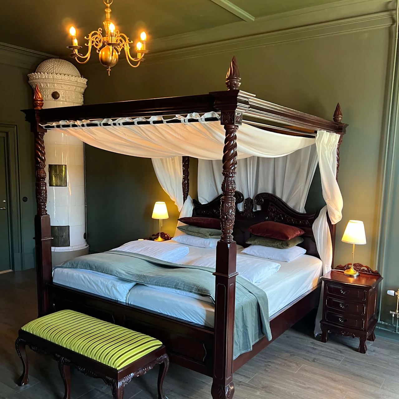 En himmelseng med hvide gardiner og grønt sengetøj står centralt i et elegant rum. En lysekrone hænger over, omgivet af grønne vægge og trægulv. Ved siden står en tændt lampe og en udsmykket bænk.