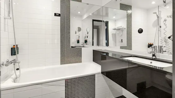 A white-tiled bathroom features a bathtub with a shower, adjacent to a sink and mirror. Context includes toiletries and a hairdryer. Black text signs read, "Water is precious, tomorrow’s shower, okay?"