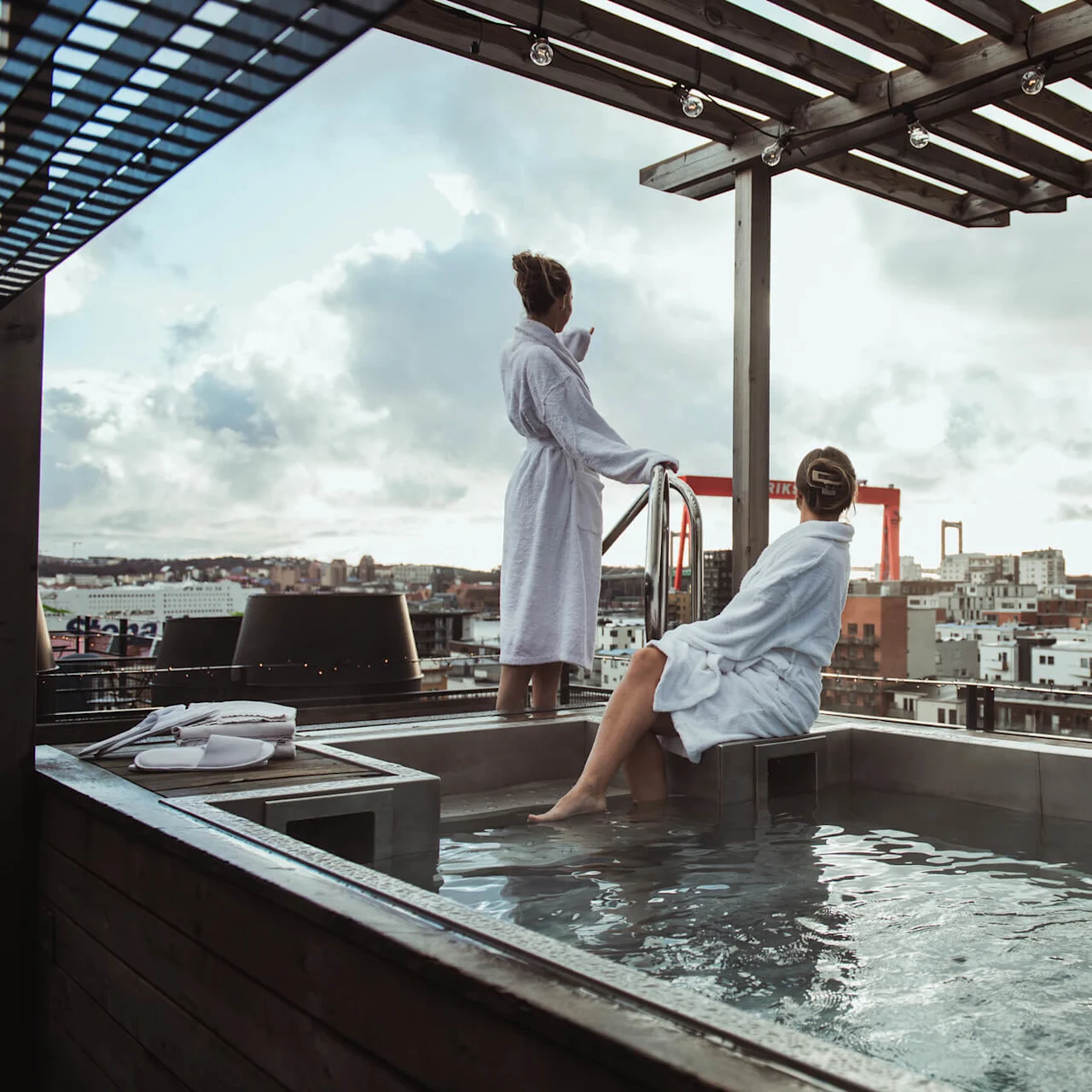 Two people in white bathrobes enjoy a rooftop pool, overlooking an urban landscape with buildings and a large crane under a cloudy sky.