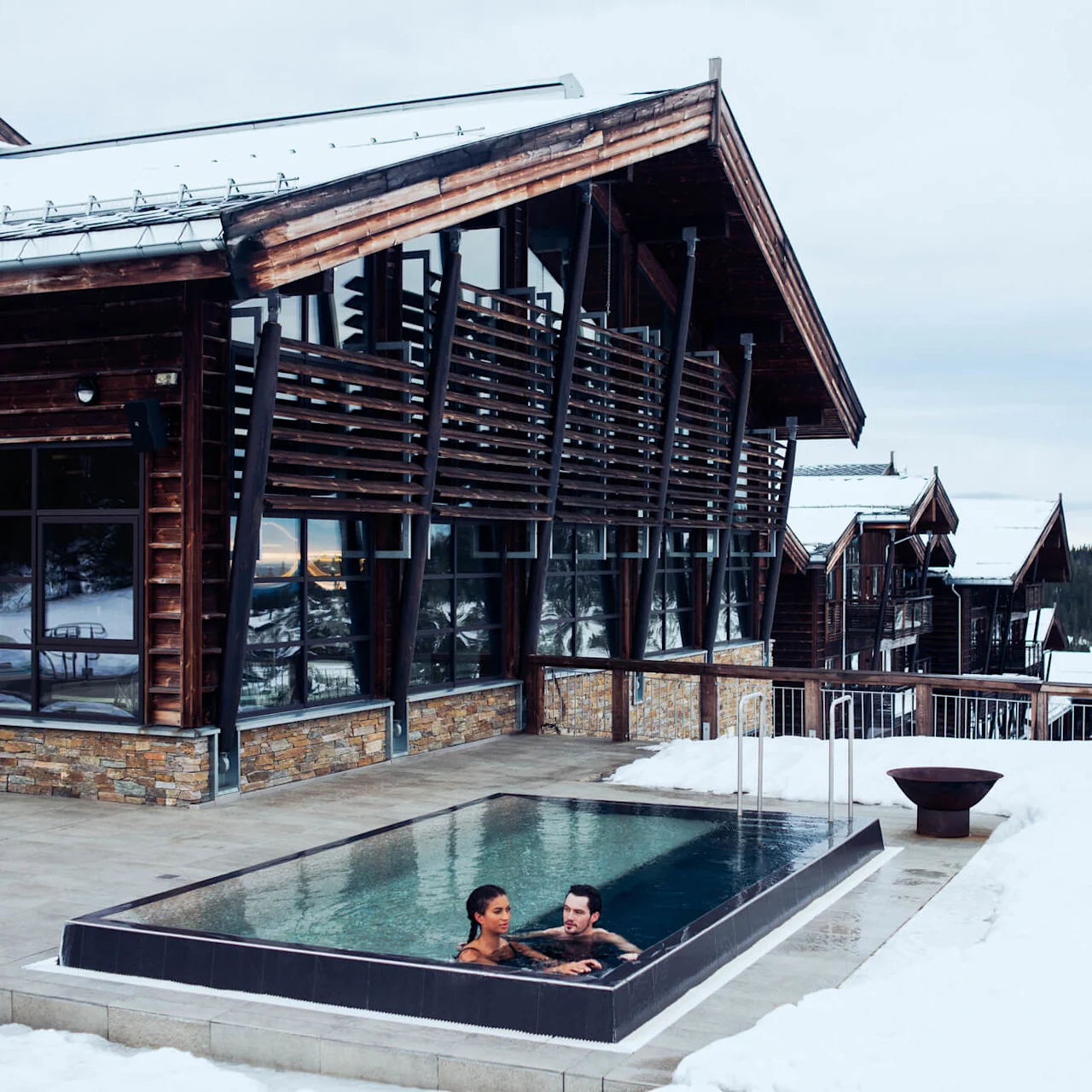 Two people relax in an outdoor heated pool beside a modern wooden lodge. Snow surrounds the area, overlooking a scenic mountain landscape.