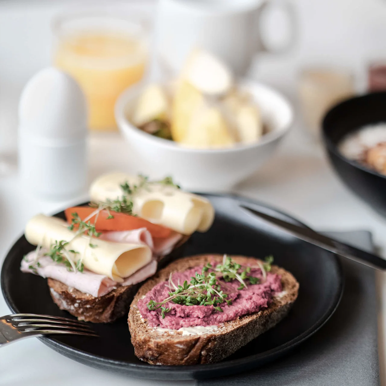 Öppna smörgåsar med kött, ost och rödbetspålägg på en svart tallrik, garnerade med örter. I bakgrunden finns en kopp, en skål med frukt och ett glas juice på ett vitt bord.