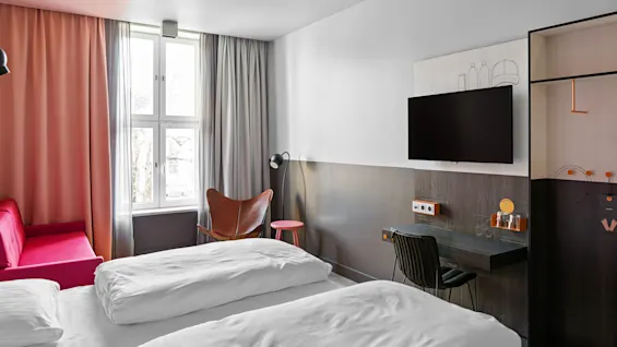 Et rent, moderne hotellrom med en dobbeltseng, rosa sofa ved et vindu med gardiner, en brun stol, en veggmontert TV og et lite skrivebord med stol.