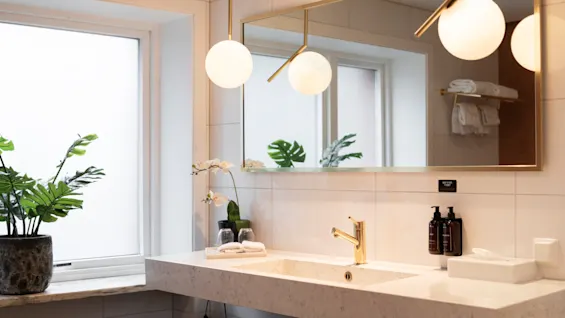 A bright and modern bathroom at Home Hotel Grand Bodø featuring a sleek sink, gold faucet, and mirror. Visible text: WAY TO SAVE WATER.