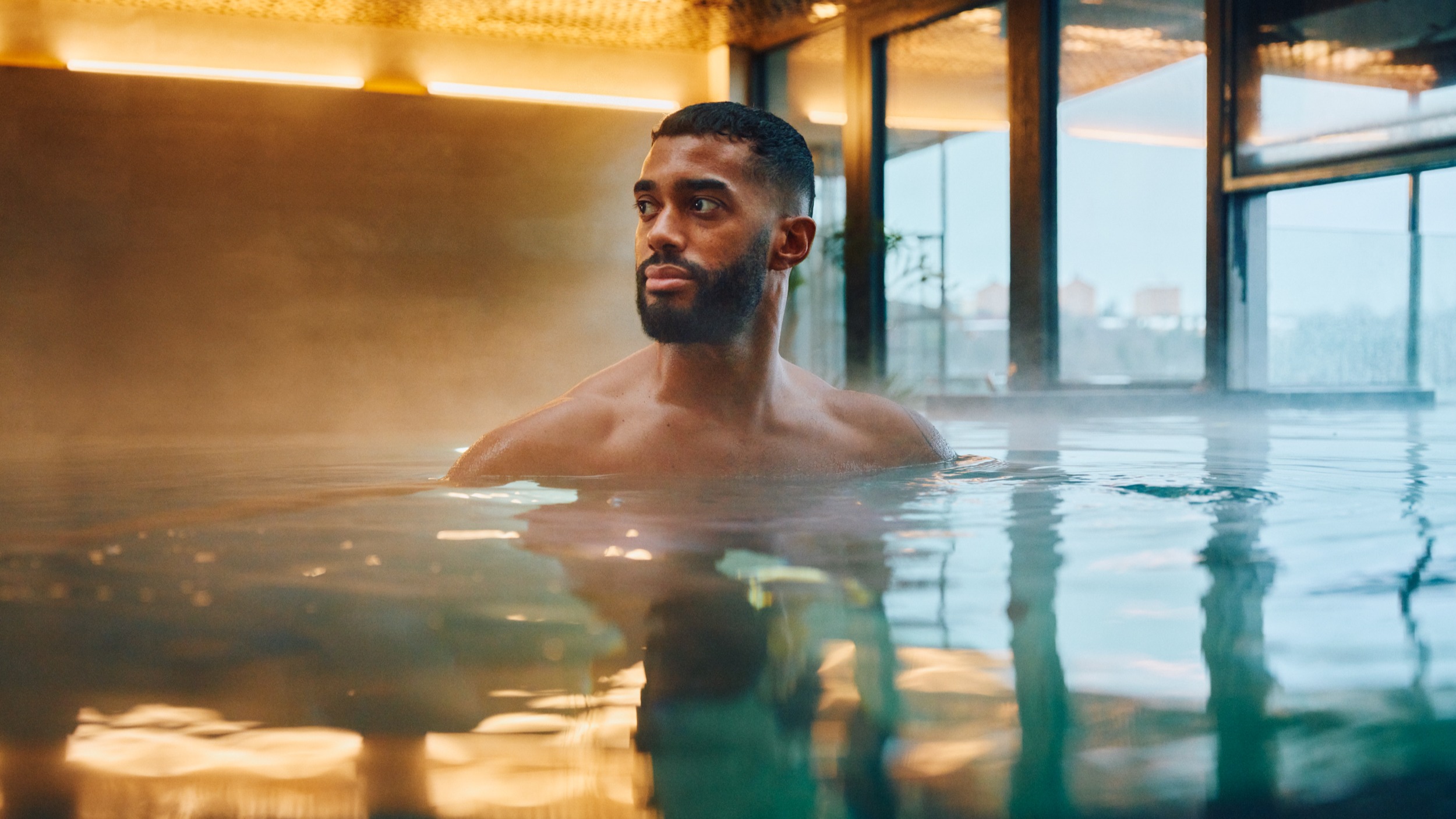 En man står i en ångande inomhuspool och tittar eftertänksamt åt sidan. Den omgivande miljön är modern med stora fönster och varm belysning.
