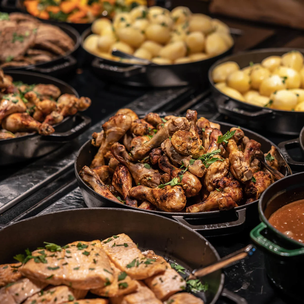 Grillede kyllingelår er arrangeret i en stor pande, omgivet af kogte kartofler, skiveskåret kød, sovs og stegte grøntsager på en buffet.