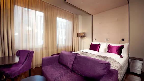 Et koselig hotellrom med en stor seng, lilla sofa, og skrivebord ved et vindu med lette gardiner.