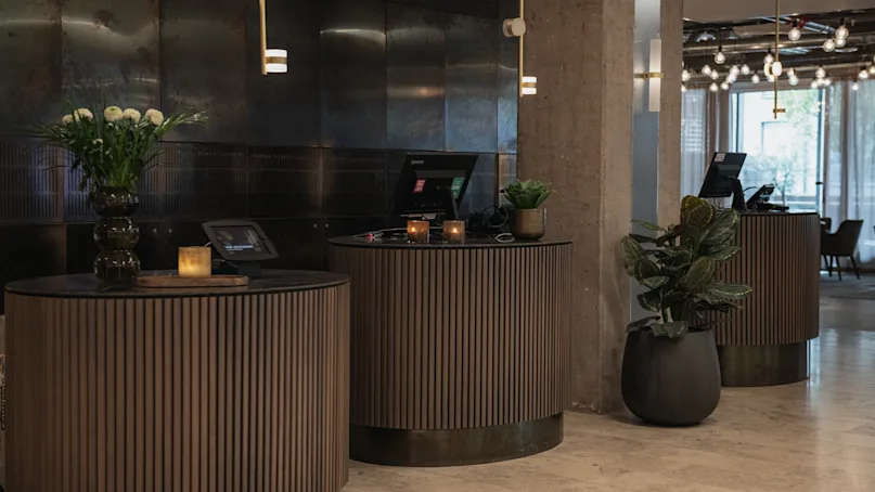 Three round wooden reception desks with computer monitors and decorative plants, positioned in a modern lobby featuring pendant lights, a concrete floor, and walls with a dark, metallic finish.