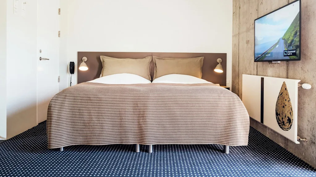 Bed under brown striped blanket, situated against a white and concrete wall with mounted TV displaying landscape and "Chromecast Built-in." Modern room with wall lamps and a blue patterned carpet.