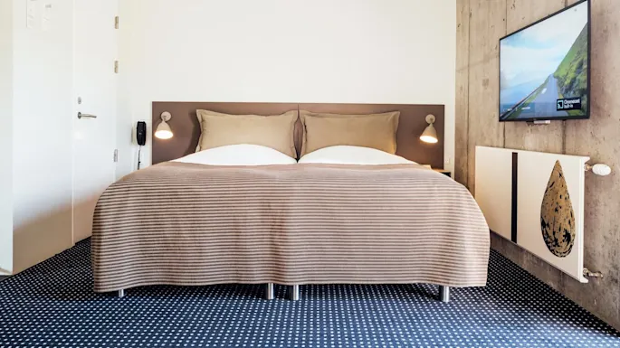 Seng under brunstripet teppe, plassert mot en hvit og betongvegg med montert TV som viser landskap og "Chromecast Built-in." Moderne rom med vegglamper og blåmønstret teppe.