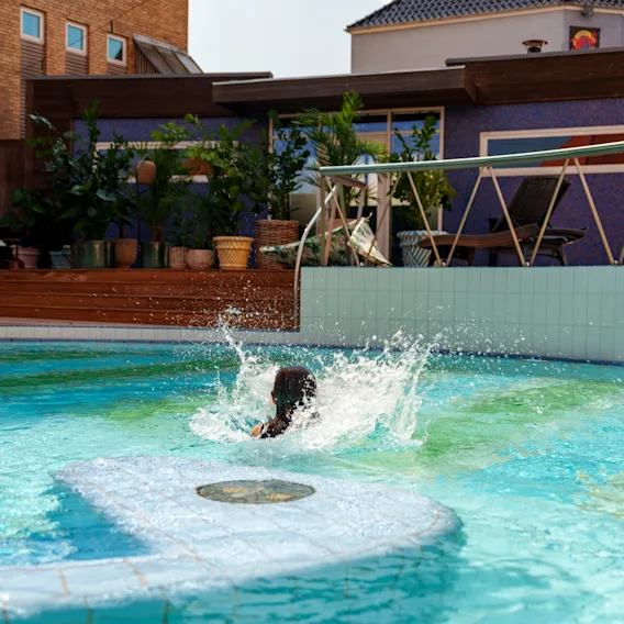 En person plaskar i en blå pool; runt dem finns krukväxter och moderna byggnader som ger en urban miljö.