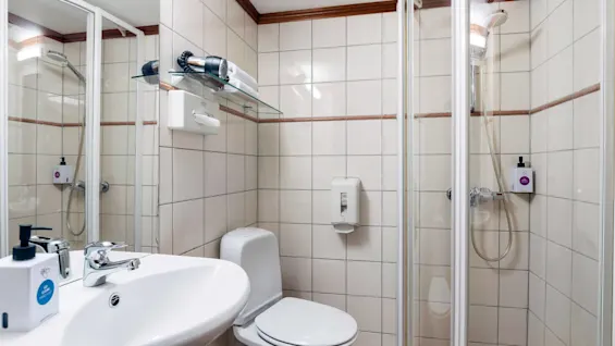 Et badeværelse med flisebelagte vægge og en brusekabine til højre. Et toilet og håndvask med vandhane i forgrunden. En hårtørrer og toiletartikler på en glashylde.