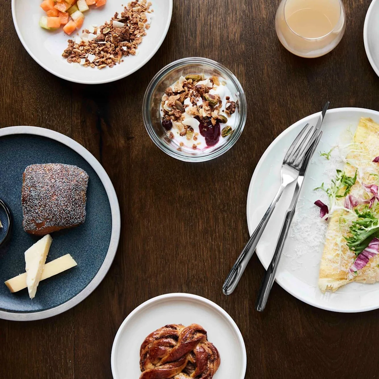 Omelette with greens sits on a plate surrounded by yogurt with granola, fruit, pastries, cheese, and coffee on a dark wooden table.