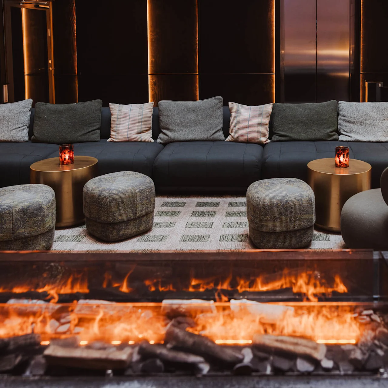 En brinnande öppen spis lyser upp en modern lounge med svarta soffor, guldiga bord med ljus och dekorativ belysning, vilket skapar en mysig atmosfär.