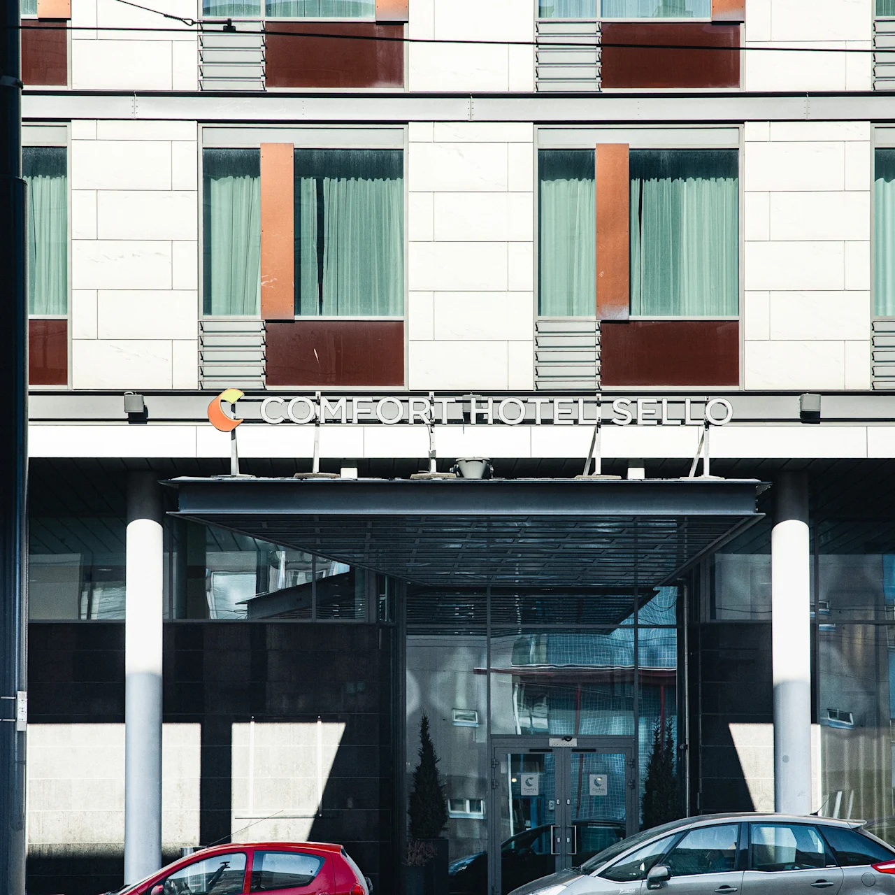 A hotel entrance is flanked by large windows and parked cars. The sign above reads "COMFORT HOTEL SELLO". Snow lines the street, hinting at a cold environment.