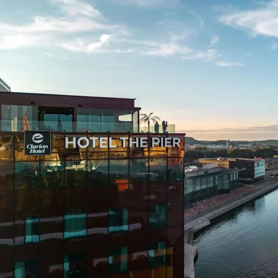 En hotellbygning merket "Clarion Hotel" og "HOTEL THE PIER" med utsikt over en elv. Takterrassen har glassrekkverk og er i et bylandskap under en blå himmel.