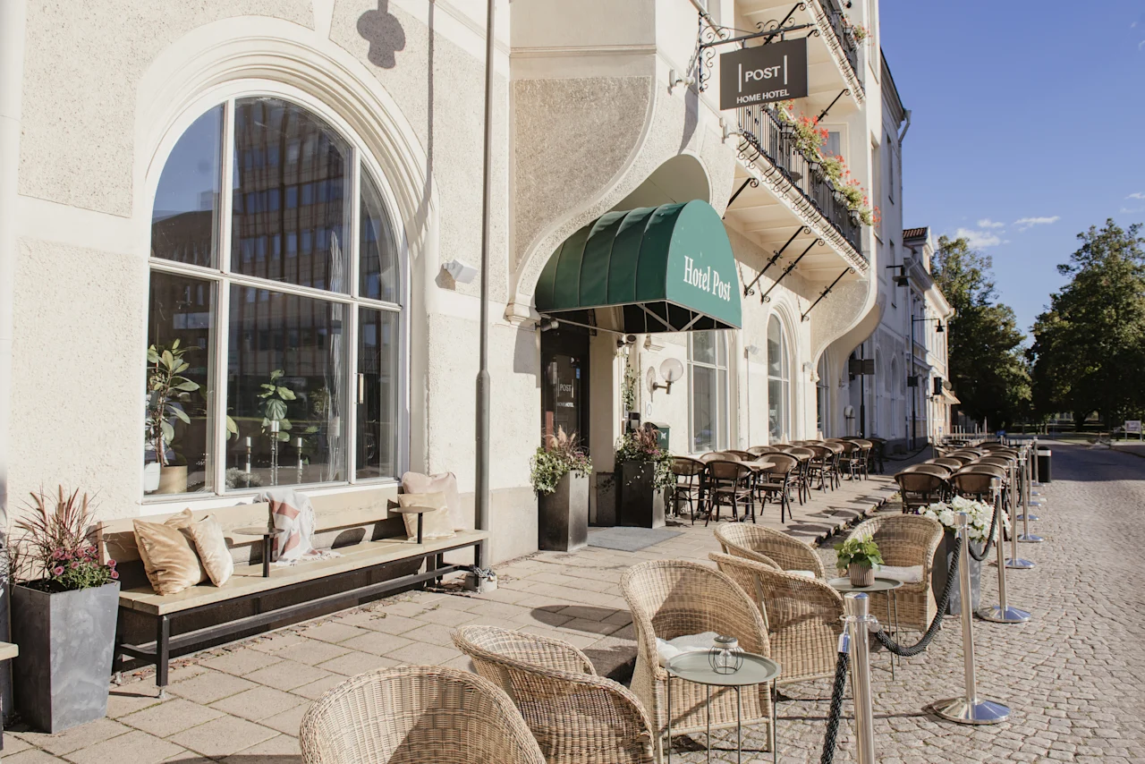 Exteriör av en byggnad med ett utomhuskafé. En skylt lyder Home Hotel Post. En markis säger Hotel Post. Flätade stolar och bord kantar trottoaren.