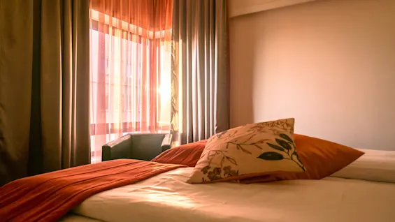 A bed is neatly made with patterned cushions and a rust-colored throw, bathed in warm sunlight streaming through sheer curtains beside a window with heavy drapes.
