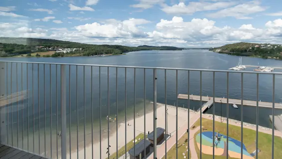 White railing overlooks peaceful waterfront with beaches and docks. Sailboats rest on calm water. Green hills with trees and clouds fill the background under a blue sky.