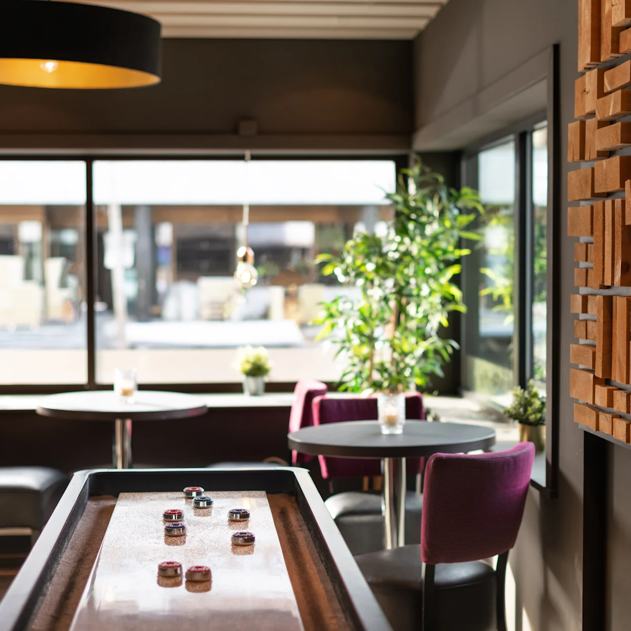 A shuffleboard table holds game pieces in a modern lounge with purple chairs and round tables by large windows, soft lighting above, and a decorative wooden wall paneling.