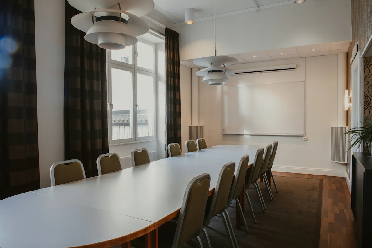 Ett väl upplyst konferensrum med ett långt bord, stolar, en whiteboard och stora fönster med mörka gardiner.