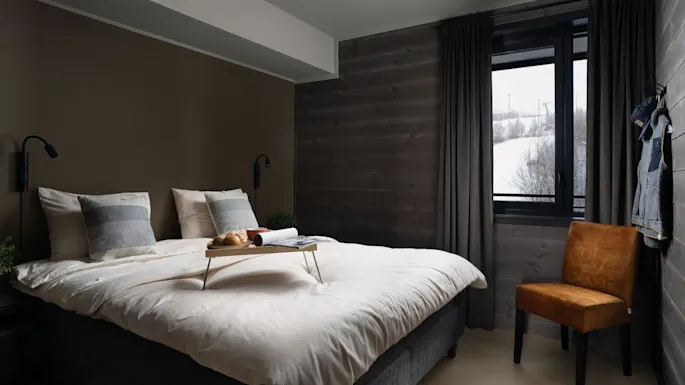 A neatly made bed with breakfast tray sits in a cozy room with brown walls and a window showing a snowy landscape. A mustard-colored chair is nearby.