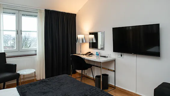 Et pænt indrettet hotelværelse med en seng, et skrivebord med stol og lampe, et fladskærms-TV på væggen, gardiner og et vindue med udsigt til træer.