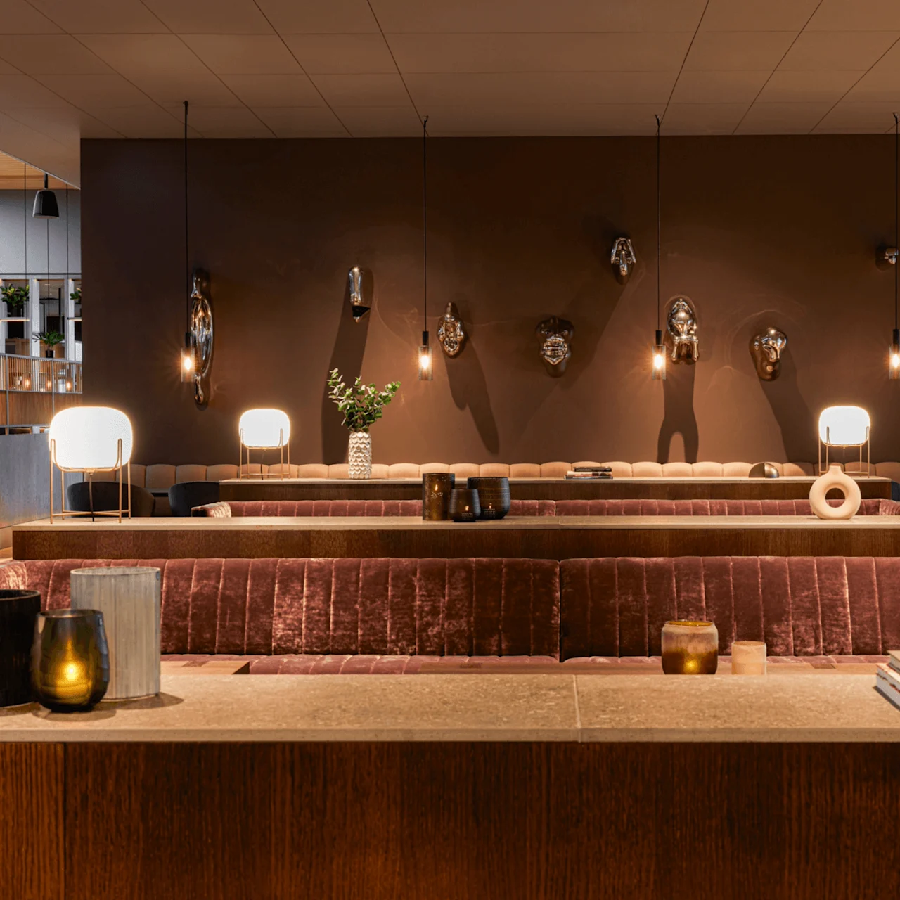 A lounge with velvet seating, decorative vases, and modern lighting. Masks adorn a brown wall. Books titled "Dion," "Object," "Out of," and "Mona Hatoum" sit on a counter. 