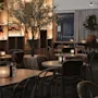 Bord med trästolar är arrangerade i en svagt upplyst restaurang med mjuka ljus, växter och ljus. En stor hylla med dekorativa föremål ger en varm och mysig atmosfär.