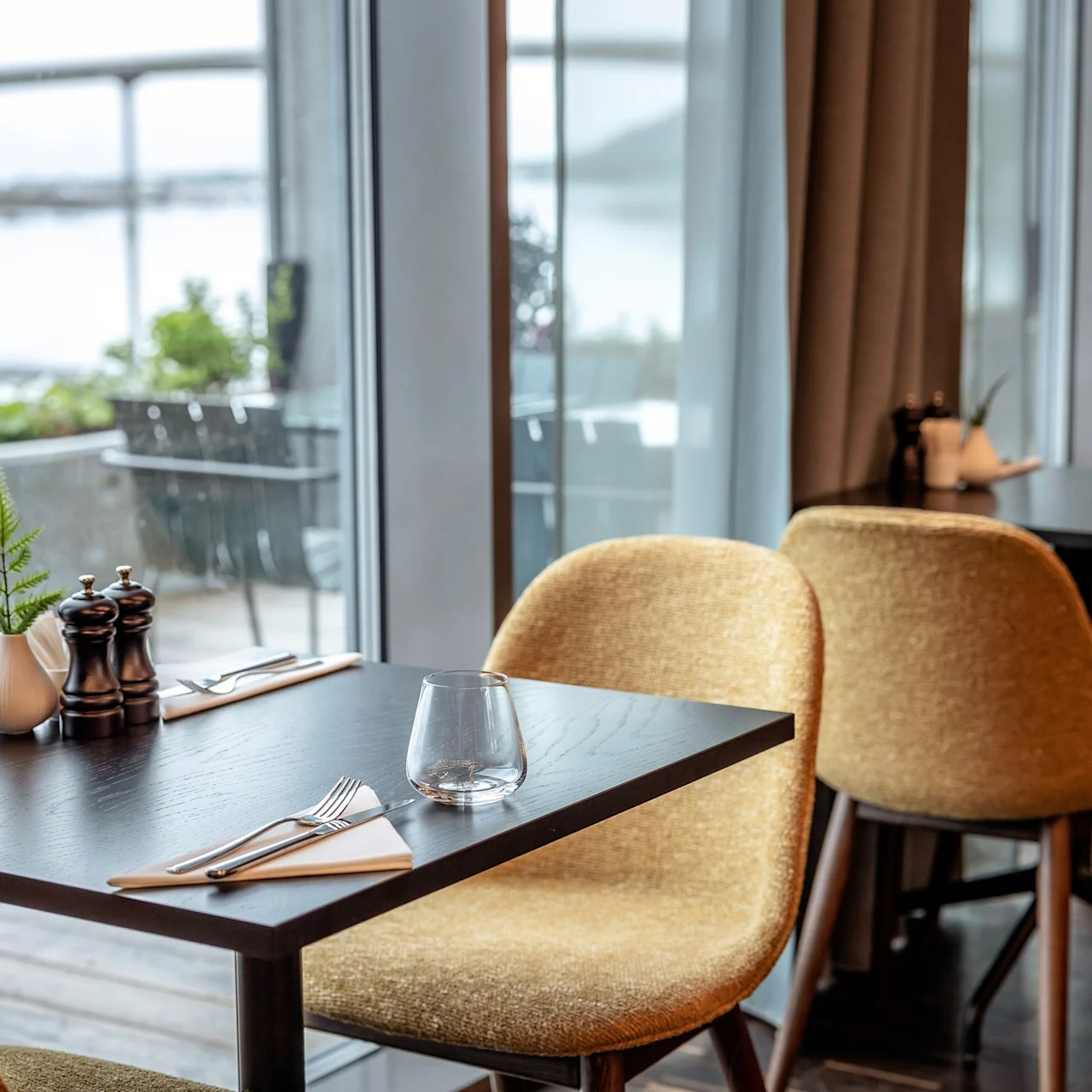 Et bord med bestik, serviet og glas er placeret ved et vindue med udsigt over en terrasse, med stole, salt- og peberbøsser og en lille plante i en moderne restaurantsammenhæng.