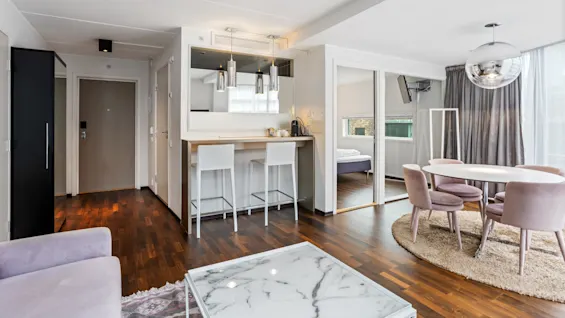 A well-lit, modern apartment interior with a connected kitchen and dining area, featuring white counters, wooden floors, and contemporary furniture.