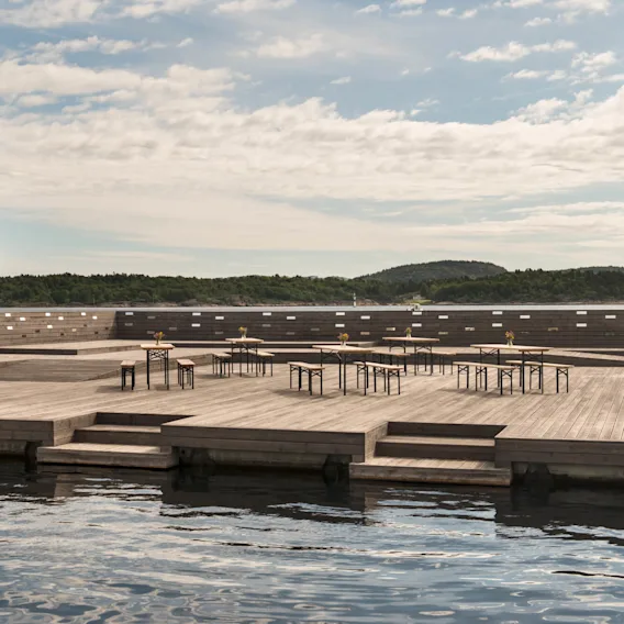 En trebygning med bord og stoler står på rolig vann, omgitt av grønne åser og bygninger, under en delvis skyet himmel.