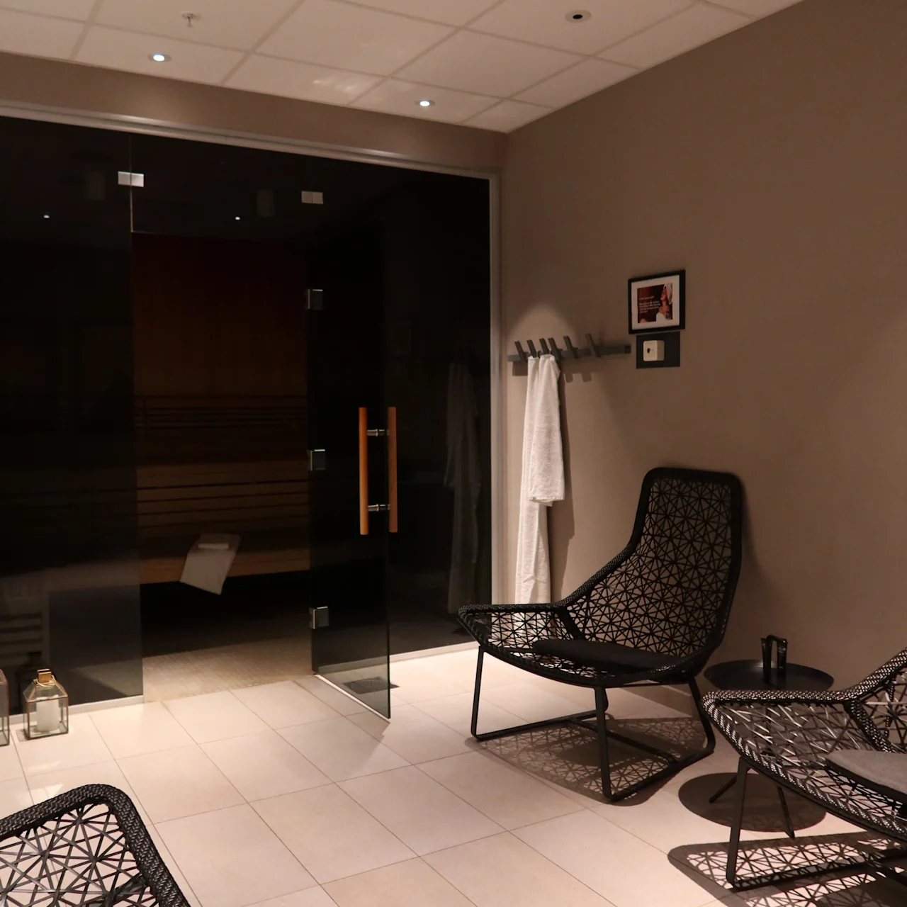 Comfortable chairs rest in a modern sauna waiting area with glass doors, soft lighting, and minimalistic decor. A coat rack holds a white robe, enhancing the relaxing spa environment.