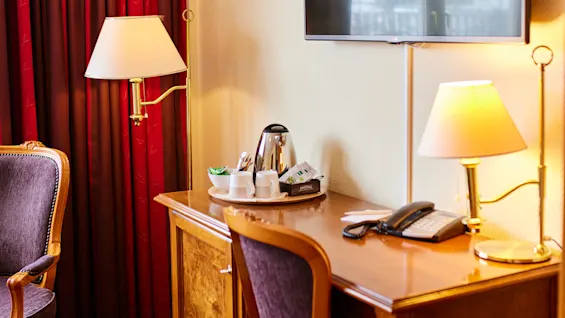 En veggmontert TV over et skrivebord i tre med et brett som inneholder en vannkoker og kopper; i nærheten, en klassisk stol og lampe med røde gardiner i et koselig hotellrom.