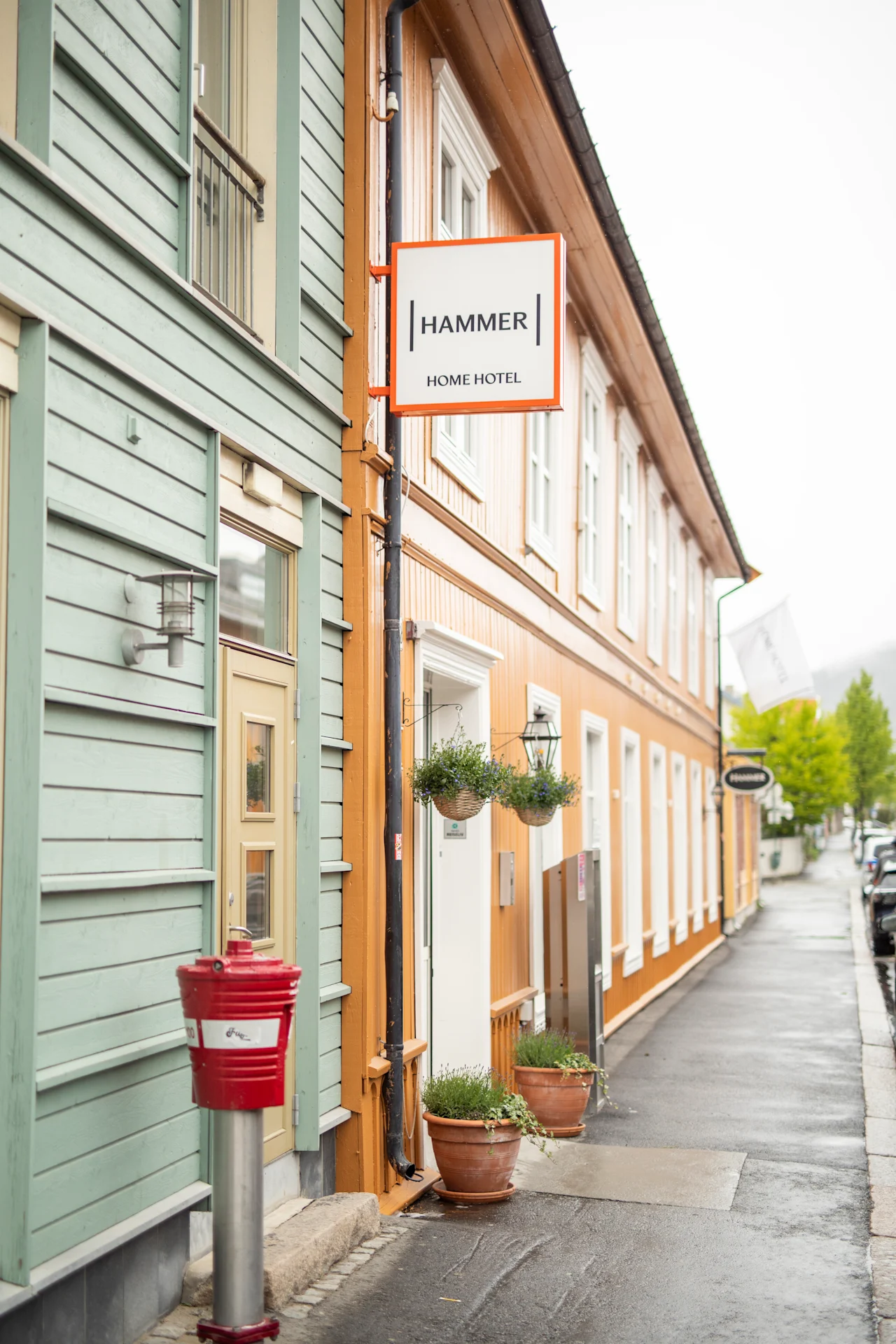A wooden building displays a sign reading "HAMMER HOME HOTEL." Hanging plants and pots decorate the entrance, and a red mailbox stands on the sidewalk beside the damp street.