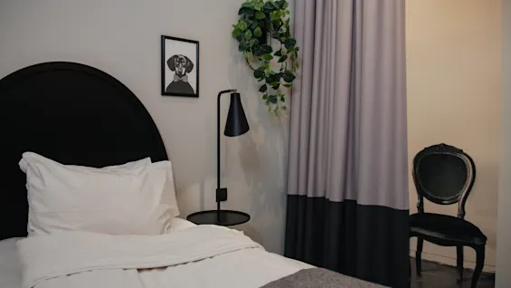 Säng med vita sängkläder i ett mysigt rum, bredvid en svart lampa och väggkonst. En stol står bakom gardiner, vilket tillför elegans till rummet.