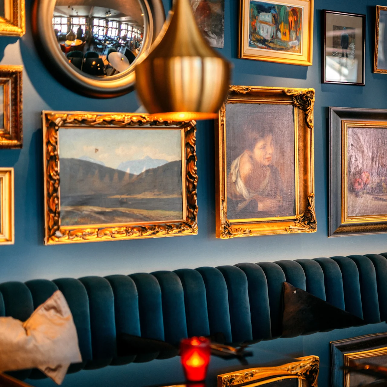 Wall adorned with framed paintings and a round mirror; beneath, a blue velvet bench with a red candle on a glossy table, creating a cozy, artful ambiance.