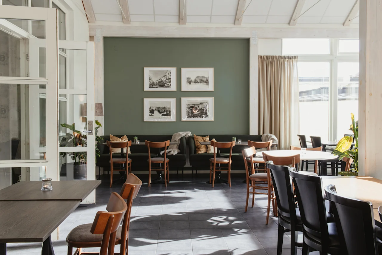 En välkomnande matplats med bord, stolar, en grön vägg med inramad konst och stora fönster. Home Hotel Post.