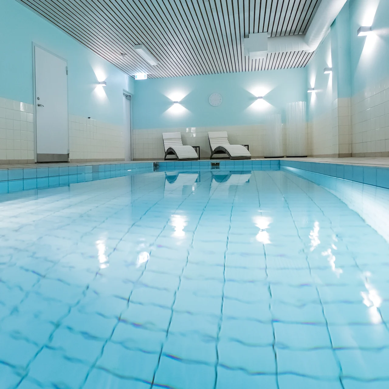 A swimming pool stretches lengthwise indoors, reflecting soft ceiling lights. Two white lounge chairs sit at the far end. The walls are painted light blue, creating a serene atmosphere.