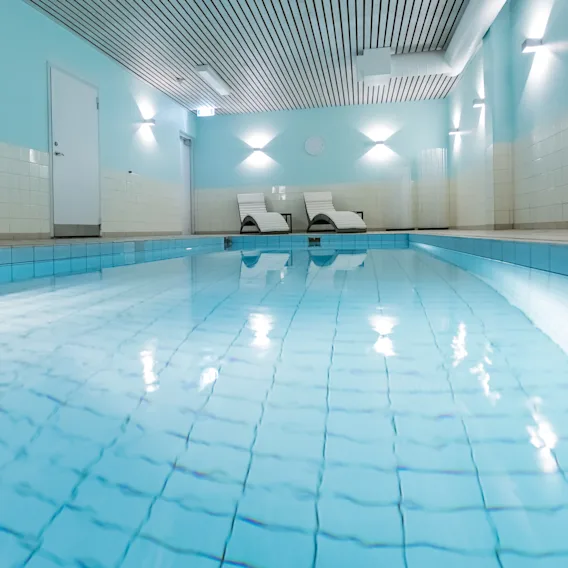 En simbassäng sträcker sig på längden inomhus och reflekterar mjuka taklampor. Två vita solstolar står i bortre änden. Väggarna är ljusblåa, vilket ger en rofylld atmosfär.
