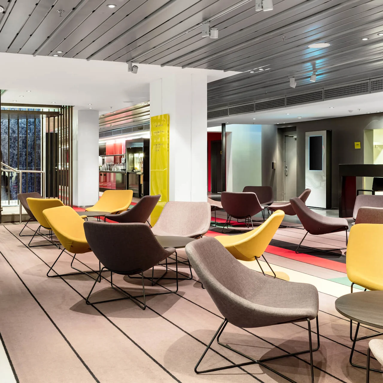 Yellow and brown lounge chairs rest on a striped carpet, surrounded by modern decor. A staircase, adorned with vertical wooden slats, leads upward. Bright, vibrant colors add to the contemporary atmosphere.
