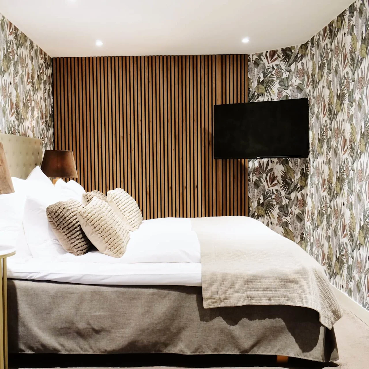 A bed with patterned cushions sits against a tropical-themed wallpaper in a cozy bedroom. A lamp rests on a side table, and a flat-screen TV is wall-mounted.