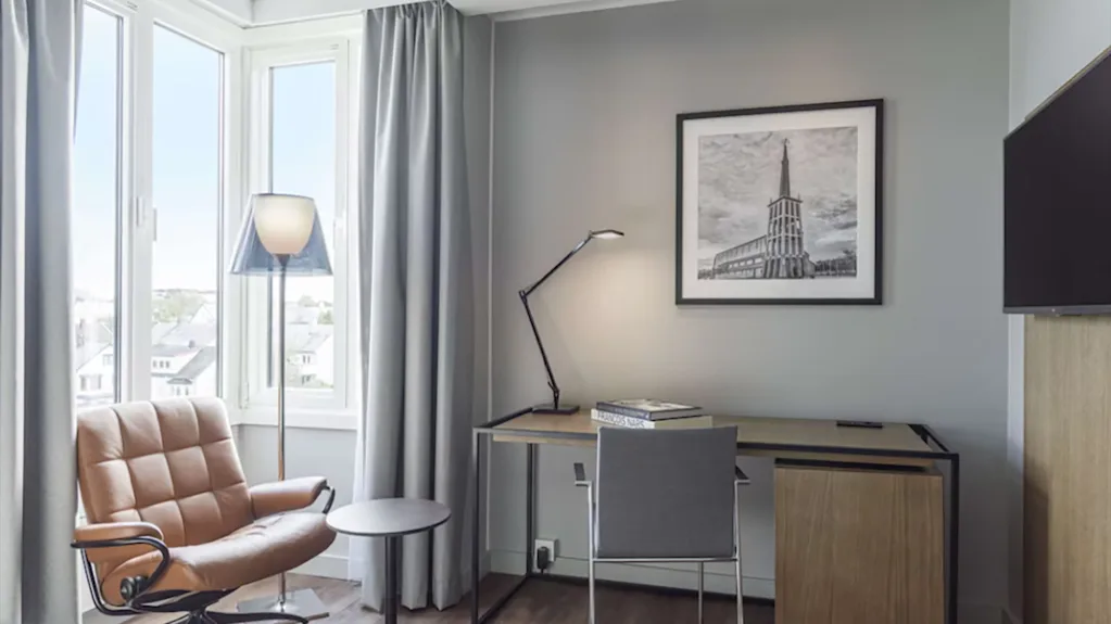 Lyst hotellrom med skrivebord, kontorstol, en behagelig skinnlenestol ved vinduet og et bilde på veggen.