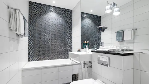Moderne badeværelse med et hvidt badekar og grå flisevæg, tilstødende håndvask og toilet. Hvide håndklæder hænger på stativer, og lys belysning skaber en ren atmosfære.
